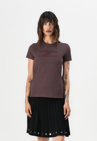 T-shirt marron à manches courtes avec un logo texturé sur le devant, associé à une jupe plissée noire avec des accents ronds en argent.