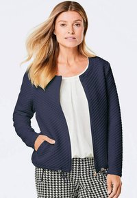 Marineblau gesteppte Jacke mit Rundhalsausschnitt und zwei Reißverschlusstaschen; getragen über einem weißen Oberteil und kombiniert mit Hahnentritt-Hosen.