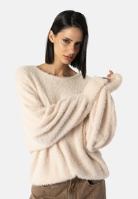 Měkký, nadýchaný svetr ve světle růžové barvě s širokým výstřihem, dlouhými rukávy a uvolněným střihem, doplněný o texturované, vysoké hnědé kalhoty.