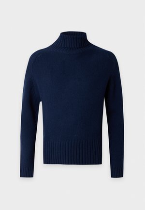 Homme portant un pull à col roulé bleu foncé, un jean bleu clair avec une ceinture marron, des chaussures marron et une casquette de baseball marron, debout devant un fond uni.