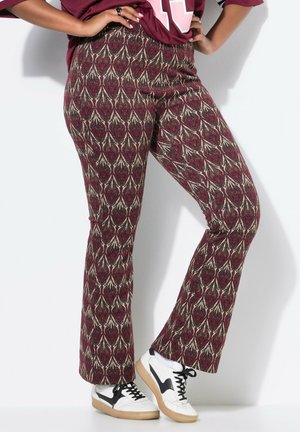 Person trägt hoch taillierte bordeauxrote gemusterte Hose und weiße Turnschuhe mit schwarzen Details, die Hände in der Hüfte, steht vor weißem Hintergrund.