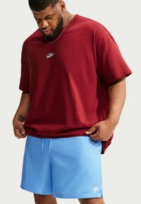 Άνδρας που φοράει κόκκινο μπλουζάκι Nike και ανοιχτό μπλε σορτς Nike κοιτάζει προς τα κάτω ενώ διορθώνει το τελείωμα της μπλούζας του.