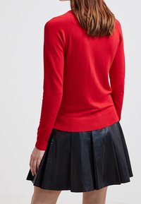 Woman wearing a red long-sleeve top and a black pleated leather skirt, standing with back facing the viewer against a plain background.