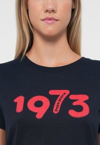 Tumši zili T-krekli no mīksta kokvilnas ar apaļu izgriezumu, uz kura ir izcili sarkanas cipars "1973" un "Pepe Jeans" modernā fontā.