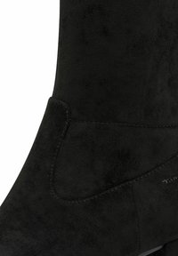 Musta mokkanahkainen nilkkuri, jossa on sileä pinta, kaareva varsi ja erottuva ompeluyksityiskohta. Varustettu pyöreällä kärjellä.