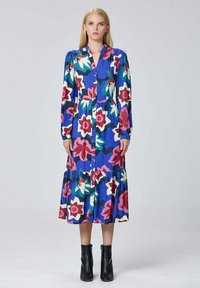 Femme se tenant debout dans une robe florale à manches longues bleu, boutonnée, ornée de fleurs roses, blanches et vertes, portant des bottines noires sur un fond uni.