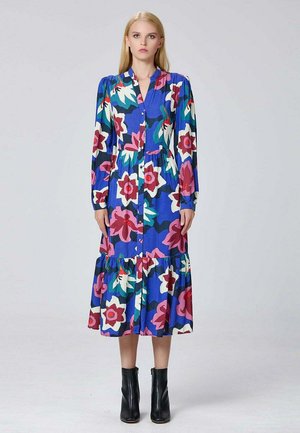 Femme se tenant debout dans une robe florale à manches longues bleu, boutonnée, ornée de fleurs roses, blanches et vertes, portant des bottines noires sur un fond uni.