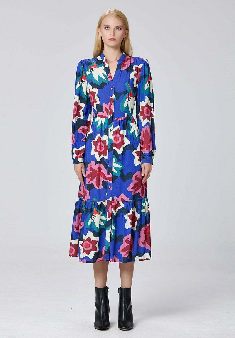 Femme se tenant debout dans une robe florale à manches longues bleu, boutonnée, ornée de fleurs roses, blanches et vertes, portant des bottines noires sur un fond uni.