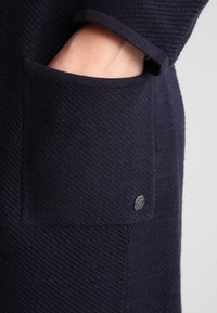 Marinblå textil med synlig ficka och metallisk logodetalj. Fickan är integrerad i en tröjddesign med ribbade kanter.