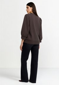 Maglione marrone con maniche a sbuffo, tessuto strutturato e scollo rotondo, abbinato a jeans neri svasati e scarpe scure.