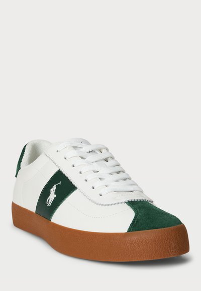 Vit sneaker med gröna mockadetaljer och ett grönt rand, med en texturerad brun gummisula, vita snören och en logotyp på sidan.
