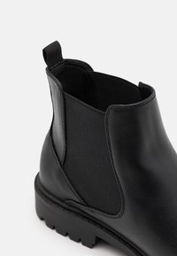 Bottine noire à tige en synthétique lisse, avec des panneaux élastiques sur les côtés et une semelle en caoutchouc robuste avec un profil texturé.