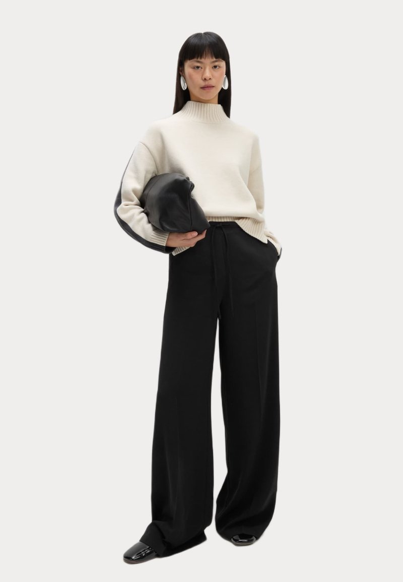 Jersey crema con cuello alto, pantalones de pierna ancha negros y un bolso de mano negro. La modelo lleva pendientes blancos llamativos, con el cabello liso y zapatos brillantes.