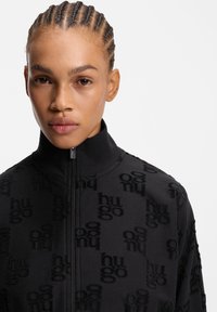 Sort lynlåsjakke med blød tekstur og præget "hu go" mønster samt høj krave. Minimalistisk design uden synligt hardware.