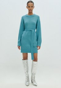 Türkisfarbenes Strickkleid mit langen Ärmeln und tailliertem Gürtel. Der Stoff wirkt weich und strukturiert. Kombiniert mit glänzenden silbernen knielangen Stiefeln.