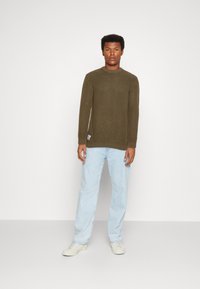 Olivgrüner, gerippter Strickpullover mit Rundhalsausschnitt, kombiniert mit hellblauen Jeans. Das Modell trägt weiße Sneaker und steht vor einem schlichten Hintergrund.