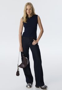 Marineblauw mouwloos ribgebreid topje gecombineerd met donkere flared jeans. Model houdt een bruine leren handtas met kettingaccenten vast. Sneakers maken de look compleet.