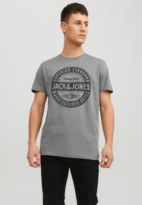 Giovane uomo che indossa una maglietta grigia Jack & Jones con un logo circolare nero, in piedi di fronte a uno sfondo chiaro e semplice, mentre guarda di lato.