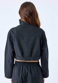 Zwarte cropped jas van textuurstof, met een hoge kraag, lange mouwen en een strakke onderrand, gecombineerd met bijpassende broek met elastische tailleband.
