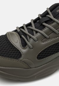 Sneaker nera e olivastro realizzata in tessuto a rete, con dettagli in pelle liscia, cuciture intricate e lacci grigi, progettata per comfort e supporto.