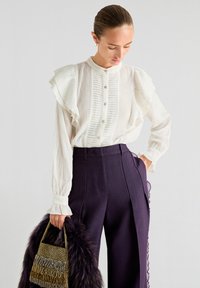 Witte, gerimpelde blouse met ribbeltextuur en knoopdetails, gecombineerd met hooggetailleerde, donkerpaarse broek met gerimpelde randen.