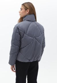 Chaqueta acolchada gris con un cuello alto, patrón acolchado y un corte relajado. Presenta puños elásticos y una silueta recortada.