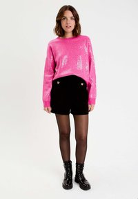 Glänzender rosa Pailletten-Pullover mit langen Ärmeln, kombiniert mit schwarzen Shorts mit herzförmigen goldenen Knöpfen und schwarzen Schnürkampfstiefeln.