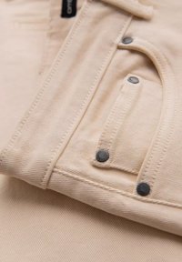 Gros plan d'une poche de veste en jean beige avec des rivets métalliques noirs visibles et des détails de couture sur un tissu texturé.