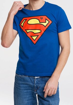 Osoba ubrana w niebieski T-shirt z dużym czerwono-żółtym logo Supermana, lekko ciągnąca lewą ręką za dekolt, ubrana w niebieskie dżinsy.