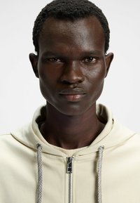 Lys beige hættebluse med en frontlynlås, struktureret stof og kontrasterende snore. Har en tætsiddende hætte og ribbedetaljer ved kraven.