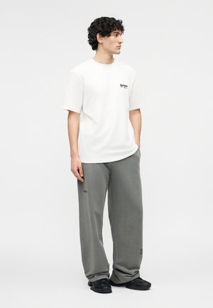 Nooruk lokkis juustega, kes kannab valget T-särki ja lahtisi halli pükse, seisab ühe käega taskus ja vaatab paremale.