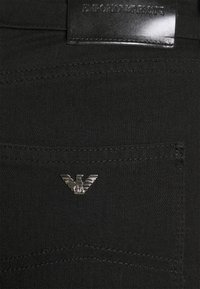 Svart denimtyg med en slät textur, med en silverfärgad metalllogga på framsidan av fickan och en svart läderetikett i midjan.