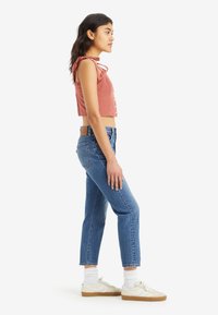 Světle růžový cropped top s ramenními šňůrkami, s designem na knoflíky; spárován s modrými rovnými jeansy a bílými teniskami s béžovými akcenty.