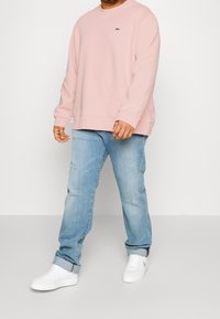 Felpa rosa chiaro con polsini e orlo a costine, abbinata a jeans azzurri e sneakers bianche. La felpa presenta un piccolo logo verde.