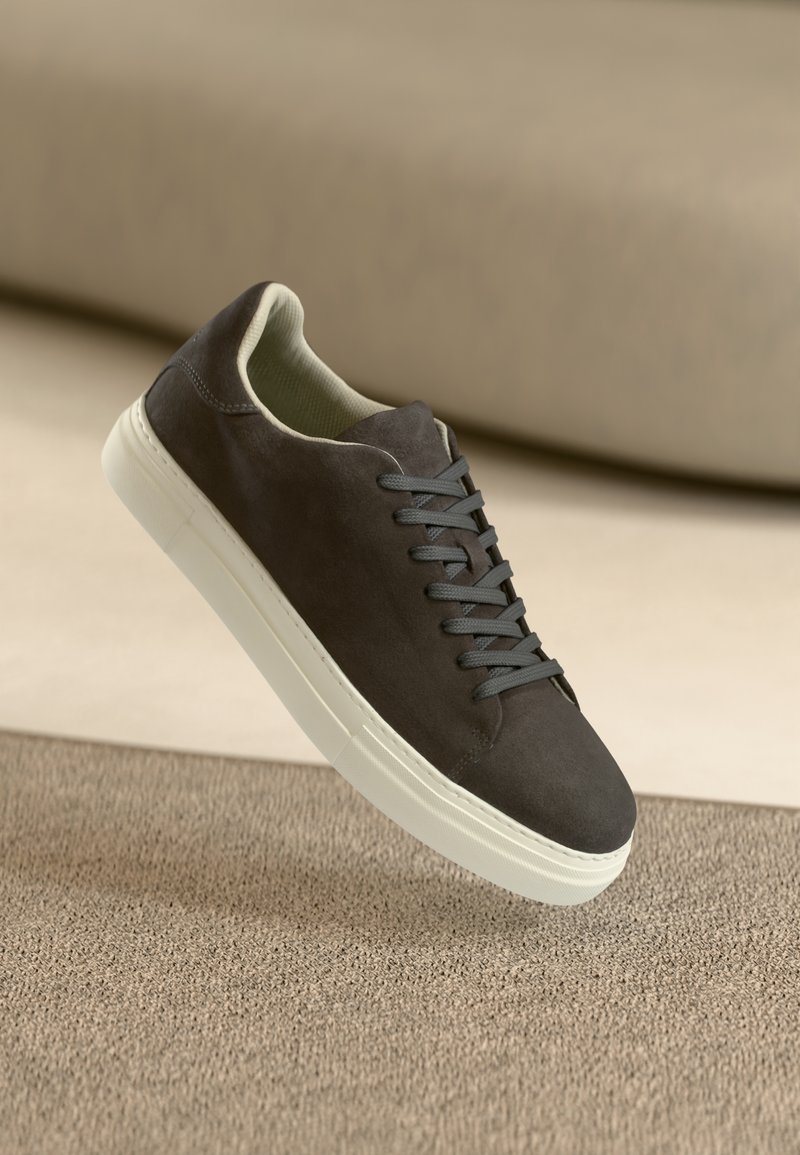 Mørkegrå semsket sneakers med matchende snørebånd og hvit gummisåle, plassert over en teksturert beige overflate med myk bakgrunn.