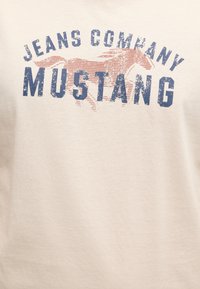 Tissu beige avec un texte bleu délavé « JEANS COMPANY MUSTANG » sur un graphisme de cheval mustang courant couleur rouille.