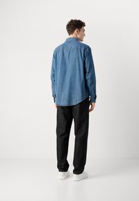 Camicia di jeans azzurra, taglio rilassato con orlo curvo, abbinata a pantaloni scuri e sneakers bianche, vista da dietro.