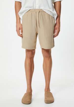 Pantaloni corti beige realizzati in un tessuto testurizzato, con vita elastica e cordoncino, tasche laterali e una linea casual. Indossati con ciabatte color sabbia.