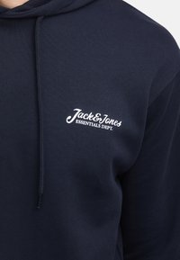 Granatowa bluza z kapturem z regulowanym sznurkiem, zakończeniami w formie ściągaczy oraz białym logo "Jack & Jones ESSENTIALS DEPT." na klatce piersiowej. Miękka w dotyku.