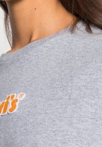 Lähikuva naisen kaulasta ja olkapäästä, joka pukeutuu harmaaseen paitaan, jossa näkyy osa oranssista ja valkoisesta brodeeratusta logosta.