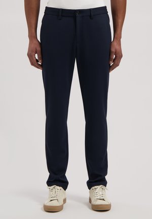 Man draagt slanke donkerblauwe broek en witte sneakers, staand tegen een effen lichte achtergrond.