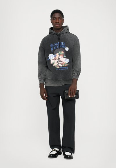 Man wearing dark gray graphic hoodie with animal print, black trousers, white and black loafers, holding a small black clutch bag.
