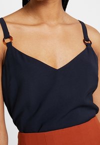 Námořnický top bez rukávů s V-výstřihem a tenkými ramínky s kulatými hnědými akcenty, vyrobený z hladkého materiálu s volným střihem.