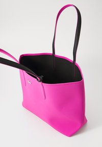 Roze tote bag met textuur op de buitenkant, open bovenkant, zwarte voering en contrasterende zwarte handvatten. Bevat een klein logo aan de zijkant.