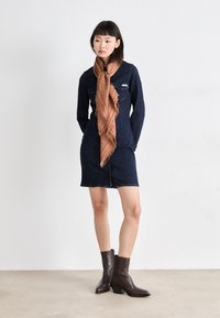 Denimkjole med lange ermer, frontlommer og knappelukking. Kombinert med et brunt mønstret skjerf og mørkebrune ankelstøvler.