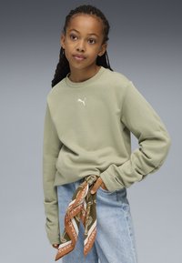 Sudadera verde claro de manga larga y cuello redondo, con un pequeño logo blanco. Combinada con jeans azules claros y una bufanda estampada.