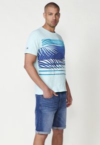 Ljusblå kortärmad t-shirt med tryck av palmblad i blå nyanser, tillsammans med uppvikta denimshorts. Avslappnad, ledig passform.