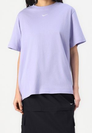 Kobieta nosząca luźny lawendowy t-shirt z krótkim rękawem z małym białym logo Nike swoosh, zestawiony z czarną spódnicą z małym logo.