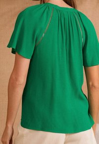 Blusa verde a maniche corte con scollatura rotonda e dettaglio arricciato sul retro. Presenta un accento testurizzato lungo le cuciture delle spalle.