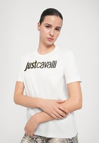 Valge lühikeste varrukate T-särk, mille esiküljel on julge, tekstureeritud musta ja kuldse "justcavalli" graafika. Pehme ja sile kangas.